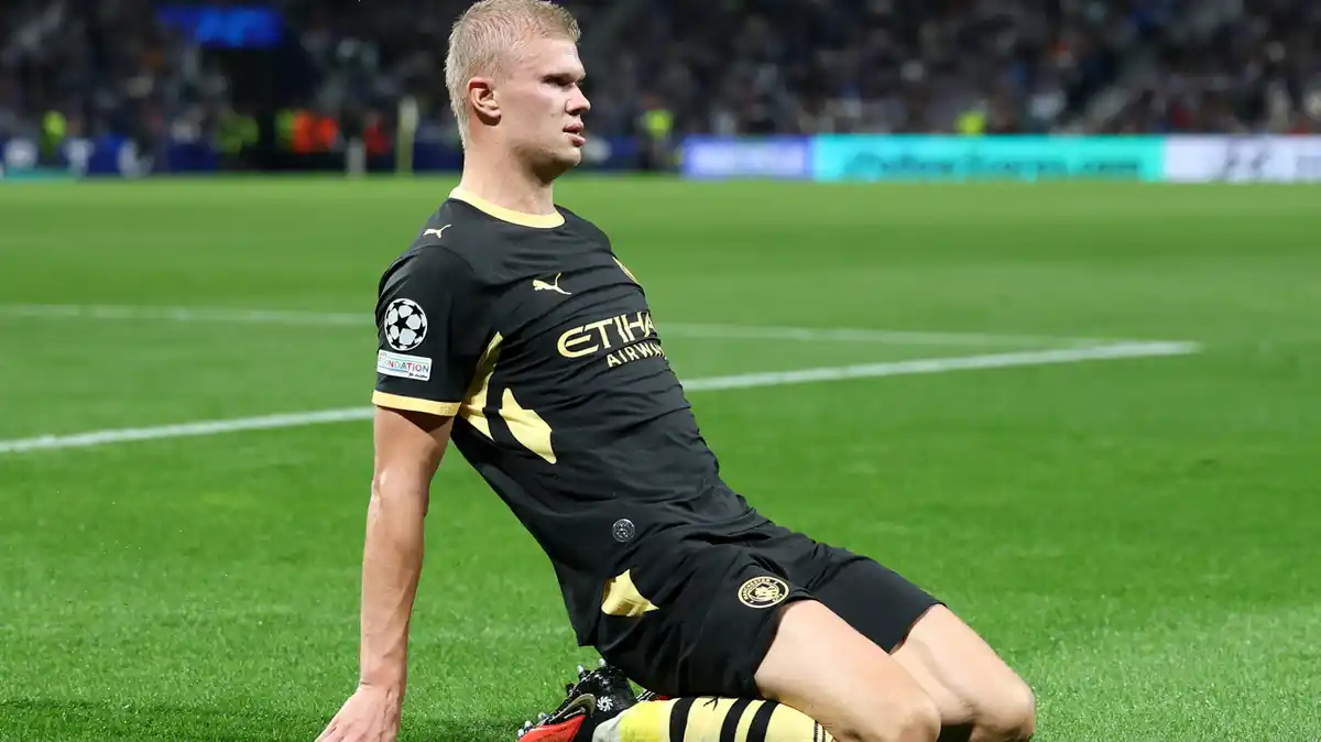Erling Haaland, wearing Manchester City's black away kit and yellow-and-black socks, slides on the turf in celebration