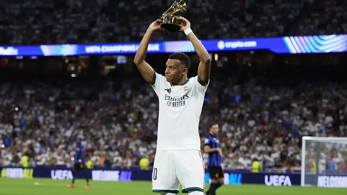 Kylian Mbappe, in Real Madrid's all-white kit, holds the Champions League top scorer trophy above his head with the Bernabeu crowd in the background