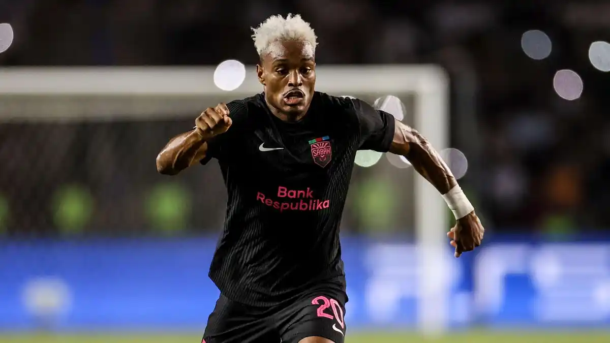 Joy-Lance Mickels, wearing an all-black Sabah kit with bright pink trim and number 20 on his shorts, spreads his arms wide as he is captured mid-stride during a football match. His hair is dyed white and he has a strange look of extreme effort on his face