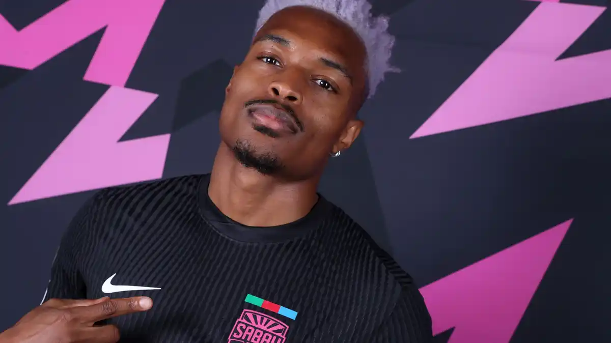 Joy-Lance Mickels, with hair dyed a light pink colour and styled extravagantly by being pushed high above his head but shaved entirely at the sides, is visible from chest up wearing a black Sabah shirt. He points with his right index finger to the bright pink club badge which reads 'Sabah' on his left breast as he looks into the camera. He stands against a black background bearing pink zigzag patterns reminiscent of lightning bolts
