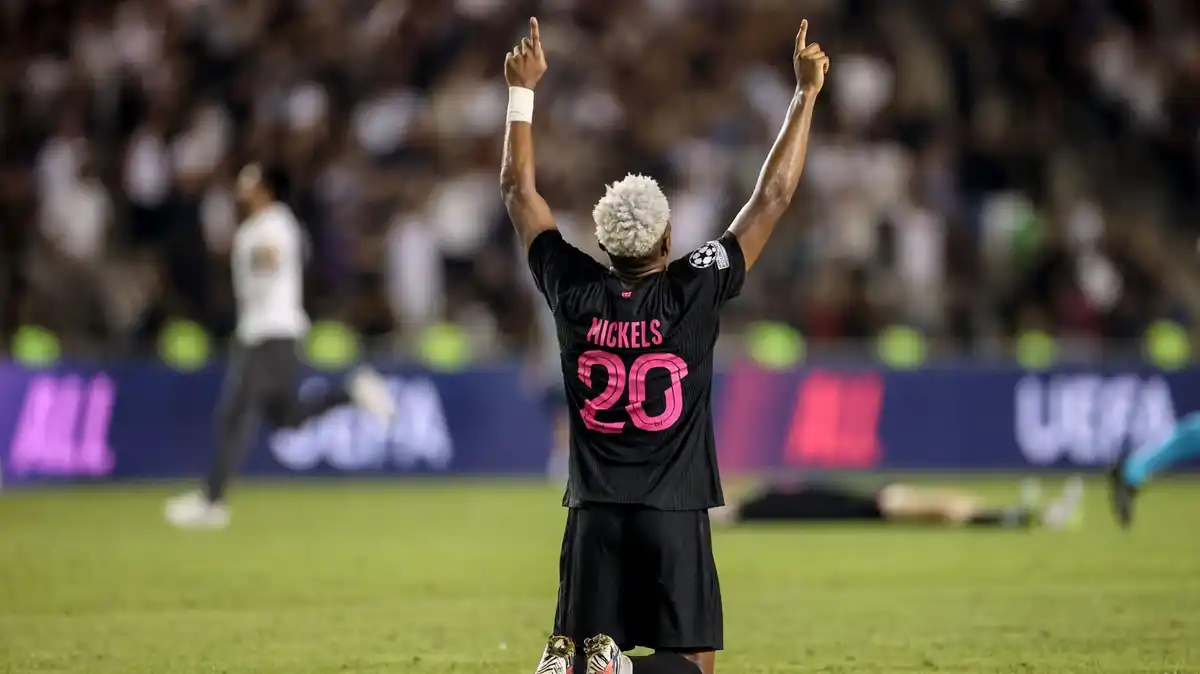 Joy-Lance Mickels, wearing an all-black Sabah kit with his surname and the number 20 in bright pink on the back, kneels down and points both hands up to the sky. The hair on the back of his head, which faces the camera, is dyed white. A full stand can be seen out of focus in the background along with two other players, with one lying on the turf and another wheeling away in celebration