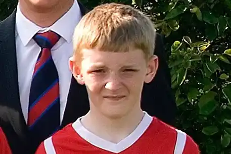 A young boy of around 14, with short blonde hair, stands looking into the camera wearing a red football kit