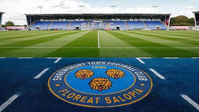 The Shrewsbury Town club crest with a stand at its home ground in the background